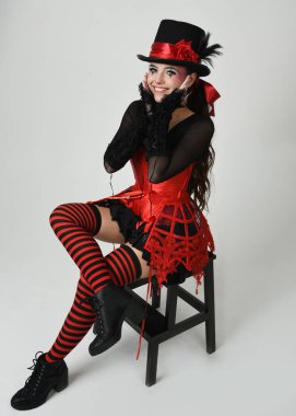 Burlesque sirk sanatçısı, Cadılar Bayramı soytarısı kostümlü fantezi kadın model silindir şapkalı, kırmızı gotik korse. Sandalyeye oturmuş tiyatro pozu, izole edilmiş beyaz stüdyo arka planı..