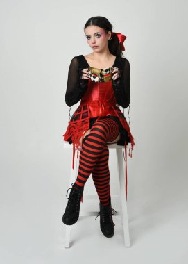 Güzel bir sirk sanatçısı, cadılar bayramı soytarısı kostümlü fantezi kadın model kırmızı gotik korse ve maskeli. Sandalyeye oturmuş tiyatro pozu, izole edilmiş beyaz stüdyo arka planı..