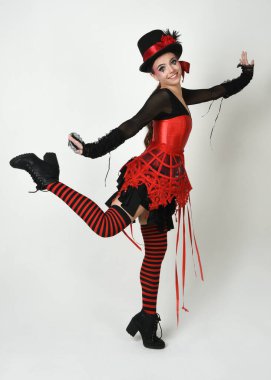 Uzun boylu, burlesk sirk sanatçısı, cadılar bayramı palyaço kostümlü, gotik şapkalı kadın model. Teatral yürüyüş pozu beyaz stüdyo arka planında izole.