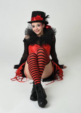 Burlesque sirk sanatçısı, Cadılar Bayramı soytarısı kostümlü fantezi kadın model silindir şapkalı, kırmızı gotik korse. Teatral oturma pozisyonu, yerde diz çökme, izole edilmiş beyaz stüdyo arka planı.