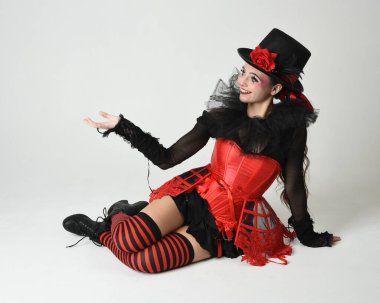 Burlesque sirk sanatçısı, Cadılar Bayramı soytarısı kostümlü fantezi kadın model silindir şapkalı, kırmızı gotik korse. Teatral oturma pozisyonu, yerde diz çökme, izole edilmiş beyaz stüdyo arka planı.