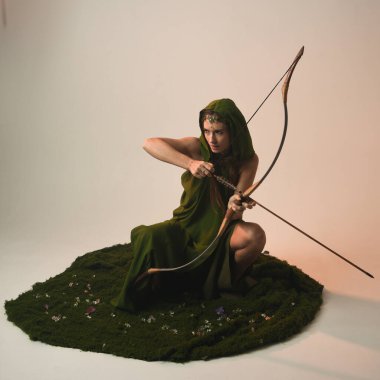 Yeşil fantezi Fae kapüşonlu başlıklı başlık kostümlü, elinde av pozunda okçuluk silahı tutan kadın modelin tam boy portresi. Yosunlu çiçekli izole stüdyo.