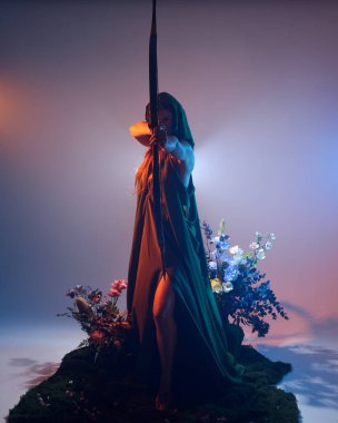 Yeşil fantezi Fae pelerini giyen, av pozisyonunda okçuluk silahı tutan, sinema renkli silüet ışıklandırmalı, yosunlu çiçeklerin arka planında duran kadın modelin dramatik bir portresi.. 