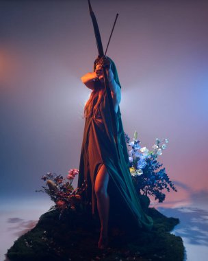 Yeşil fantezi Fae pelerini giyen, av pozisyonunda okçuluk silahı tutan, sinema renkli silüet ışıklandırmalı, yosunlu çiçeklerin arka planında duran kadın modelin dramatik bir portresi.. 