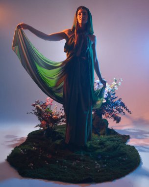 Yeşil fantezi elf pelerin ve akıcı kumaşlı dramatik bir kadın modeli portresi. Yosunlu orman arka planında yürüyen kişinin renkli arka ışıklandırmalı silueti..