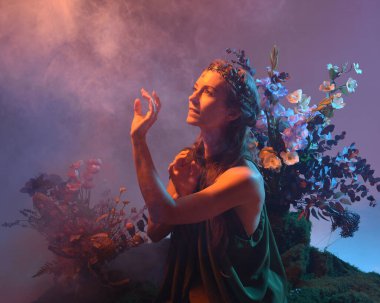 Yeşil peri elbisesi giymiş güzel bir kadın modelin dramatik fantezi portresi. Çiçeklerle çevrili, yosun kaplı sandalyede otururken kol hareketi uzanıyor. arka ışıklandırmalı siluetli figür, karamsar ışıklandırma stüdyosu arka planı.