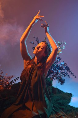 Yeşil peri elbisesi giymiş güzel bir kadın modelin dramatik fantezi portresi. Çiçeklerle çevrili, yosun kaplı sandalyede otururken kol hareketi uzanıyor. arka ışıklandırmalı siluetli figür, karamsar ışıklandırma stüdyosu arka planı.