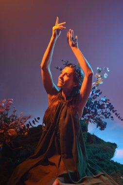 Yeşil peri elbisesi giymiş güzel bir kadın modelin dramatik fantezi portresi. Çiçeklerle çevrili, yosun kaplı sandalyede otururken kol hareketi uzanıyor. arka ışıklandırmalı siluetli figür, karamsar ışıklandırma stüdyosu arka planı.