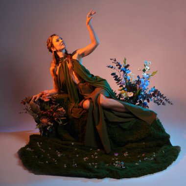 Dramatik, uzun, fantezi portresi güzel kadın model yeşil peri elbisesi giyiyor. Doğa çiçekleriyle çevrili yosun kaplı sandalyede oturuyorlar. Altın günbatımı silueti figürü, stüdyo arka planı.