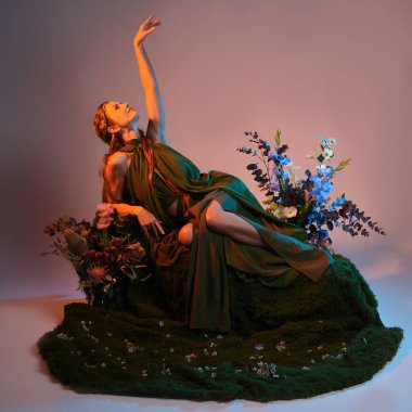 Dramatik, uzun, fantezi portresi güzel kadın model yeşil peri elbisesi giyiyor. Doğa çiçekleriyle çevrili yosun kaplı sandalyede oturuyorlar. Altın günbatımı silueti figürü, stüdyo arka planı.