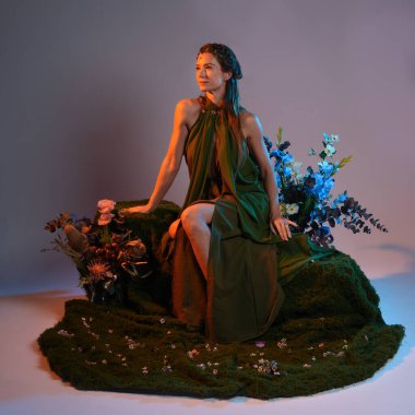 Dramatik, uzun, fantezi portresi güzel kadın model yeşil peri elbisesi giyiyor. Doğa çiçekleriyle çevrili yosun kaplı sandalyede oturuyorlar. Altın günbatımı silueti figürü, stüdyo arka planı.