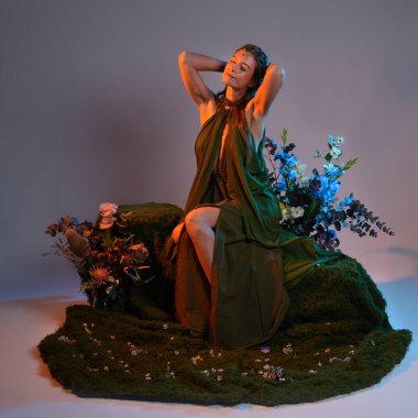 Dramatik, uzun, fantezi portresi güzel kadın model yeşil peri elbisesi giyiyor. Doğa çiçekleriyle çevrili yosun kaplı sandalyede oturuyorlar. Altın günbatımı silueti figürü, stüdyo arka planı.