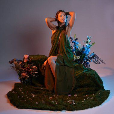 Dramatik, uzun, fantezi portresi güzel kadın model yeşil peri elbisesi giyiyor. Doğa çiçekleriyle çevrili yosun kaplı sandalyede oturuyorlar. Altın günbatımı silueti figürü, stüdyo arka planı.