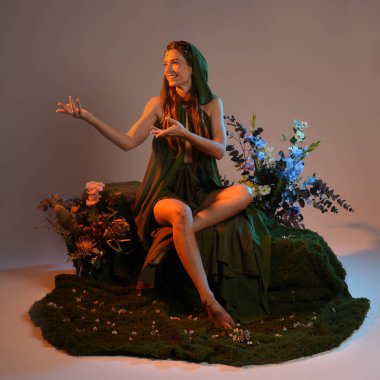Dramatik, uzun, fantezi portresi güzel kadın model yeşil peri elbisesi giyiyor. Doğa çiçekleriyle çevrili yosun kaplı sandalyede oturuyorlar. Altın günbatımı silueti figürü, stüdyo arka planı.