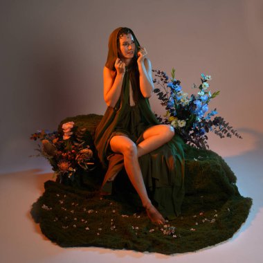 Dramatik, uzun, fantezi portresi güzel kadın model yeşil peri elbisesi giyiyor. Doğa çiçekleriyle çevrili yosun kaplı sandalyede oturuyorlar. Altın günbatımı silueti figürü, stüdyo arka planı.