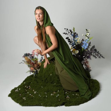 Dramatik fantezi portresi güzel kadın model yeşil peri elbisesi giyiyor. Çiçeklerle çevrili yosun kaplı sandalyede otururken. Stüdyo arkaplanındaki izole kişi.