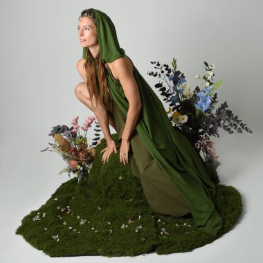 Dramatik fantezi portresi güzel kadın model yeşil peri elbisesi giyiyor. Çiçeklerle çevrili yosun kaplı sandalyede otururken. Stüdyo arkaplanındaki izole kişi.