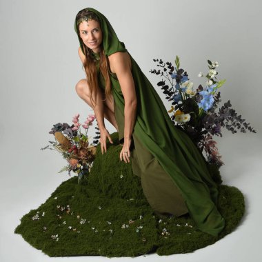 Dramatik fantezi portresi güzel kadın model yeşil peri elbisesi giyiyor. Çiçeklerle çevrili yosun kaplı sandalyede otururken. Stüdyo arkaplanındaki izole kişi.
