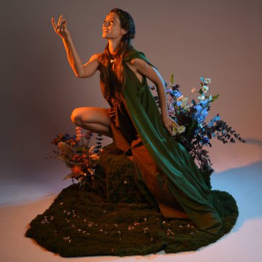 Dramatik, uzun, fantezi portresi güzel kadın model yeşil peri elbisesi giyiyor. Doğa çiçekleriyle çevrili yosun kaplı sandalyede oturuyorlar. Altın günbatımı silueti figürü, stüdyo arka planı.