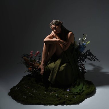 Yeşil peri elbisesi ve taçlı pelerin giyen güzel esmer kadın modelin dramatik fantezi portresi. Çiçeklerle çevrili yosun kaplı sandalyede otururken. arka planda siluetli bir figür, stüdyo arka planında karamsar ışıklandırma.