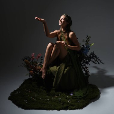 Yeşil peri elbisesi ve taçlı pelerin giyen güzel esmer kadın modelin dramatik fantezi portresi. Çiçeklerle çevrili yosun kaplı sandalyede otururken. arka planda siluetli bir figür, stüdyo arka planında karamsar ışıklandırma.