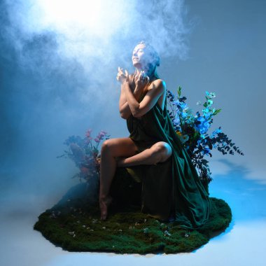 Yeşil peri elbisesi ve taçlı pelerin giyen güzel esmer kadın modelin dramatik fantezi portresi. Çiçeklerle çevrili yosun kaplı sandalyede otururken. Stüdyo arka planında renkli ay ışığı olan sisli duman efektleri.