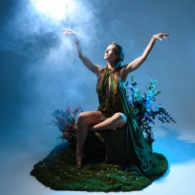 Yeşil peri elbisesi ve taçlı pelerin giyen güzel esmer kadın modelin dramatik fantezi portresi. Çiçeklerle çevrili yosun kaplı sandalyede otururken. Stüdyo arka planında renkli ay ışığı olan sisli duman efektleri.