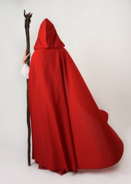 Ortaçağ kırmızı kapüşonlu fantezi pelerini giyen tam boy bir insan figürü. Kameradan uzaklaşmak, geriye bakmak, sihirbaz asasını tutmak. Beyaz stüdyo arka planında izole edilmiş..