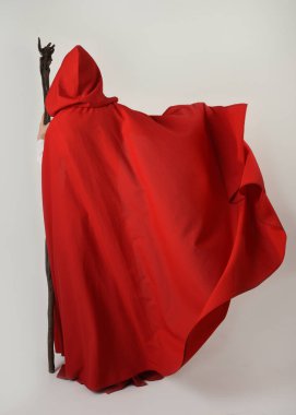 Ortaçağ kırmızı kapüşonlu fantezi pelerini giyen tam boy bir insan figürü. Kameradan uzaklaşmak, geriye bakmak, sihirbaz asasını tutmak. Beyaz stüdyo arka planında izole edilmiş..