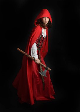 Kırmızı kapüşonlu peri masalı pelerini ve ortaçağ tarihi elbisesi giyen güzel esmer bir kadının dramatik portresi. Elinde balta var. Karanlık stüdyo arka planında izole bir figür..