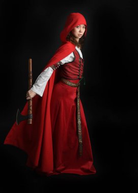 Kırmızı kapüşonlu peri masalı pelerini ve ortaçağ tarihi elbisesi giyen güzel esmer bir kadının dramatik portresi. Elinde balta var. Karanlık stüdyo arka planında izole bir figür..