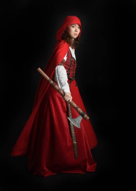 Kırmızı kapüşonlu peri masalı pelerini ve ortaçağ tarihi elbisesi giyen güzel esmer bir kadının dramatik portresi. Elinde balta var. Karanlık stüdyo arka planında izole bir figür..