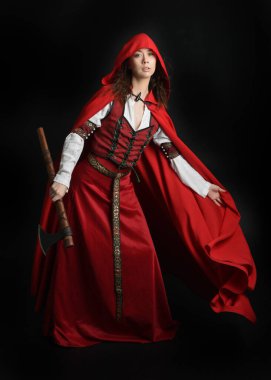 Kırmızı kapüşonlu peri masalı pelerini ve ortaçağ tarihi elbisesi giyen güzel esmer bir kadının dramatik portresi. Elinde balta var. Karanlık stüdyo arka planında izole bir figür..