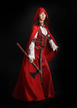 Kırmızı kapüşonlu peri masalı pelerini ve ortaçağ tarihi elbisesi giyen güzel esmer bir kadının dramatik portresi. Elinde balta var. Karanlık stüdyo arka planında izole bir figür..