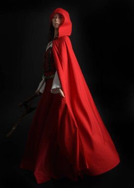 Kırmızı kapüşonlu peri masalı pelerini ve ortaçağ tarihi elbisesi giyen güzel esmer bir kadının dramatik portresi. Elinde balta var. Karanlık stüdyo arka planında izole bir figür..
