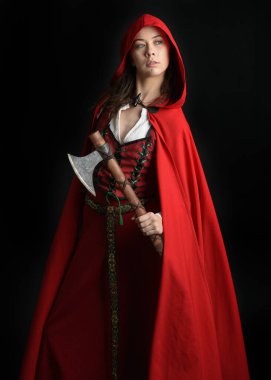 Kırmızı kapüşonlu peri masalı pelerini ve ortaçağ tarihi elbisesi giyen güzel esmer bir kadının dramatik portresi. Elinde balta var. Karanlık stüdyo arka planında izole bir figür..