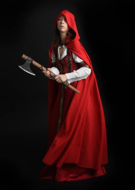 Kırmızı kapüşonlu peri masalı pelerini ve ortaçağ tarihi elbisesi giyen güzel esmer bir kadının dramatik portresi. Elinde balta var. Karanlık stüdyo arka planında izole bir figür..