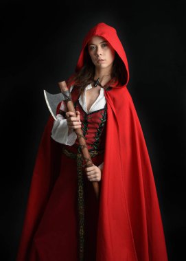 Kırmızı kapüşonlu peri masalı pelerini ve ortaçağ tarihi elbisesi giyen güzel esmer bir kadının dramatik portresi. Elinde balta var. Karanlık stüdyo arka planında izole bir figür..