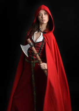 Kırmızı kapüşonlu peri masalı pelerini ve ortaçağ tarihi elbisesi giyen güzel esmer bir kadının dramatik portresi. Elinde balta var. Karanlık stüdyo arka planında izole bir figür..