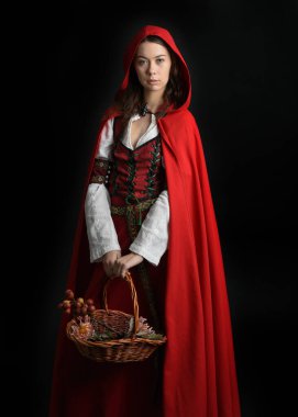 Kırmızı kapüşonlu peri masalı pelerini ve ortaçağ tarihi elbisesi giyen güzel esmer bir kadının dramatik portresi. Hasır sepeti tutuyorum. Karanlık stüdyo arka planında izole bir figür..