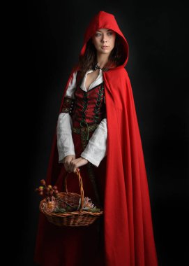 Kırmızı kapüşonlu peri masalı pelerini ve ortaçağ tarihi elbisesi giyen güzel esmer bir kadının dramatik portresi. Hasır sepeti tutuyorum. Karanlık stüdyo arka planında izole bir figür..