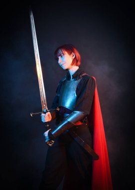 Ortaçağ tarihi zırh kostümlü kızıl saçlı fantezi savaşçı kadın ve kırmızı peri masalı pelerin, güçlü kahraman şövalyenin sinematik portresi, destansı savaş pozu, karamsar arkaplanına karşı dramatik siluet