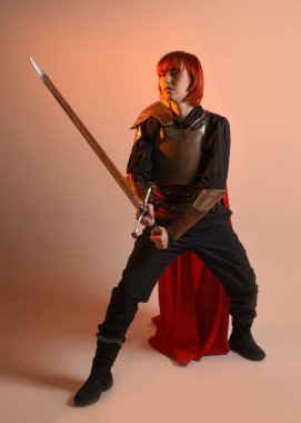 Fantezi savaşçısı kadın modelinin portresi, elinde kılıç, kırmızı peri masalı pelerinli tarihi ortaçağ zırhı, güçlü bir fantezi şövalyesi. izole edilmiş stüdyo arka planı altın sinematik ışıklandırma.