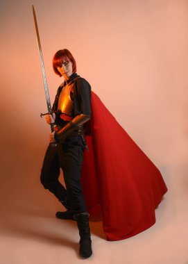 Fantezi savaşçısı kadın modelinin portresi, elinde kılıç, kırmızı peri masalı pelerinli tarihi ortaçağ zırhı, güçlü bir fantezi şövalyesi. izole edilmiş stüdyo arka planı altın sinematik ışıklandırma.