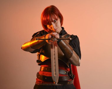 Fantezi savaşçısı kadın modelinin portresi, elinde kılıç, kırmızı peri masalı pelerinli tarihi ortaçağ zırhı, güçlü bir fantezi şövalyesi. izole edilmiş stüdyo arka planı altın sinematik ışıklandırma.