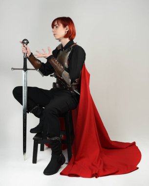 Tarihi bir ortaçağ zırhı giymiş fantezi savaşçı kadın modeli, kırmızı peri masalı pelerini, güçlü bir fantezi şövalyesinin sinematik portresi. Oturma pozisyonu kılıç, stüdyo arka planı.