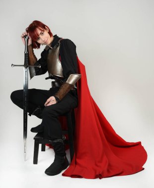 Tarihi bir ortaçağ zırhı giymiş fantezi savaşçı kadın modeli, kırmızı peri masalı pelerini, güçlü bir fantezi şövalyesinin sinematik portresi. Oturma pozisyonu kılıç, stüdyo arka planı.