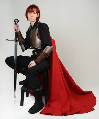 Tarihi bir ortaçağ zırhı giymiş fantezi savaşçı kadın modeli, kırmızı peri masalı pelerini, güçlü bir fantezi şövalyesinin sinematik portresi. Oturma pozisyonu kılıç, stüdyo arka planı.