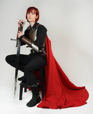 Tarihi bir ortaçağ zırhı giymiş fantezi savaşçı kadın modeli, kırmızı peri masalı pelerini, güçlü bir fantezi şövalyesinin sinematik portresi. Oturma pozisyonu kılıç, stüdyo arka planı.