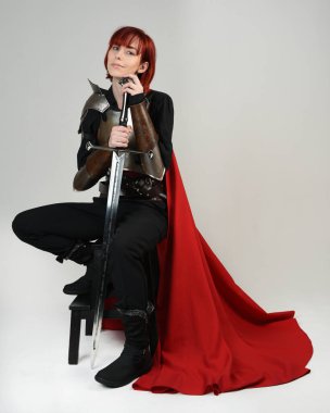 Tarihi bir ortaçağ zırhı giymiş fantezi savaşçı kadın modeli, kırmızı peri masalı pelerini, güçlü bir fantezi şövalyesinin sinematik portresi. Oturma pozisyonu kılıç, stüdyo arka planı.